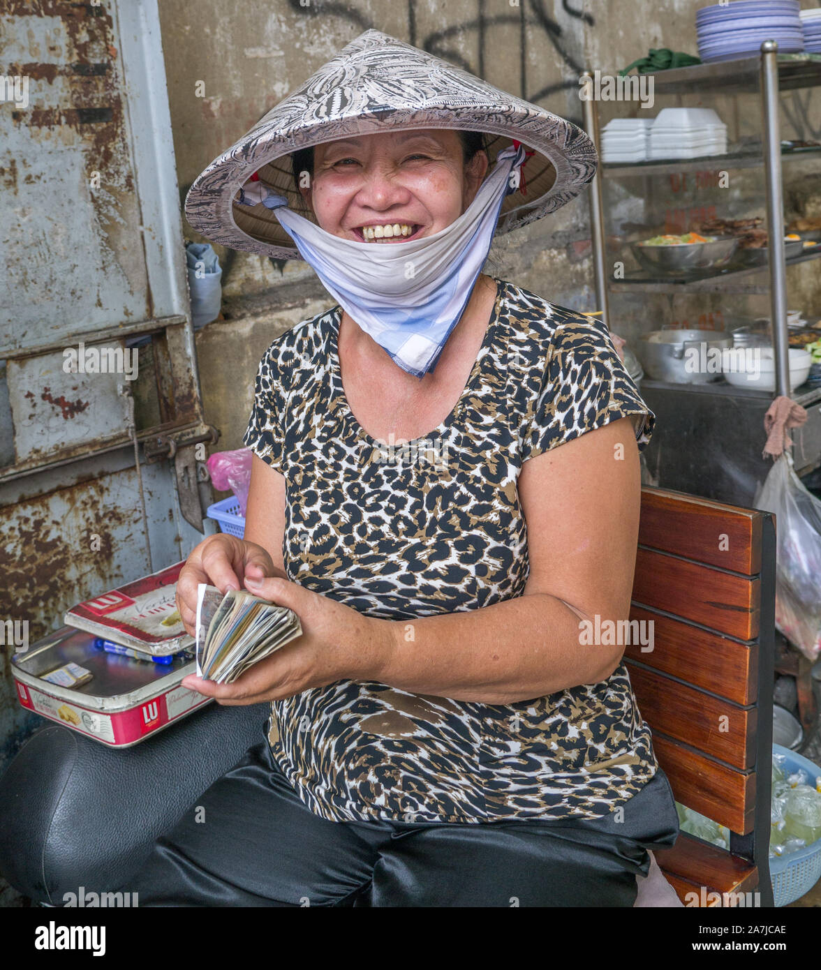 Lächelnde, vietnamesische Straßenverkäuferin, die Strohhut trägt, weißer Kinnschal auf dem Stuhl sitzt und Geld einen großen Stapel auf Geld in der rechten Hand zählt Stockfoto