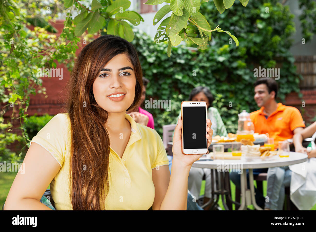 Lächelnd weibliche Mobile-Phone zeigen im Garten zu Hause mit der Familie essen Frühstück im Hintergrund Stockfoto