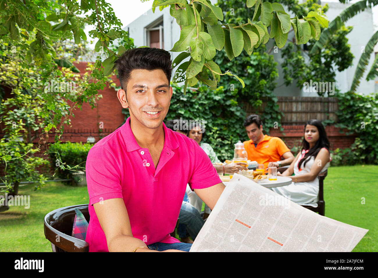 Lächelnd nach Mann-in-Stuhl und lesen Zeitung mit Familie im Hintergrund im Innenhof ihres Hauses Stockfoto