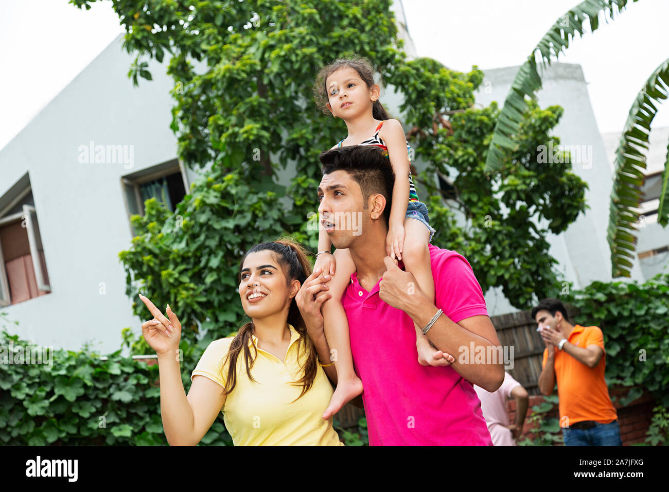 Glückliche junge Familie Mutter mit Kind Mädchen auf Schultern und Papa zeigen auf etwas im Hof in der Nähe von Ihrem Haus Stockfoto