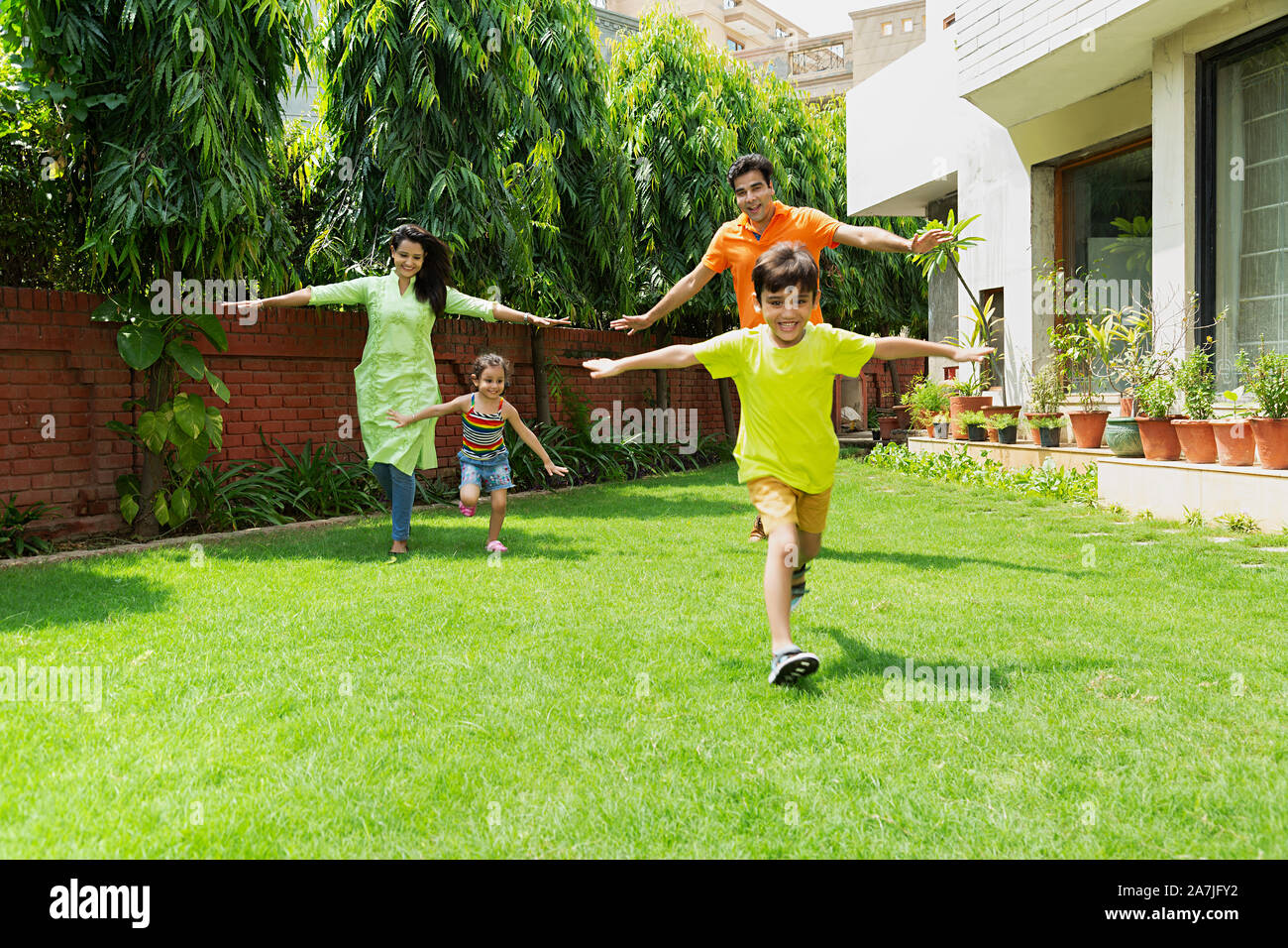 Gerne indische Familie Eltern zwei Kinder mit Arms-Outstretched In-Garden laufen zusammen in der Nähe von Ihrem Haus Stockfoto