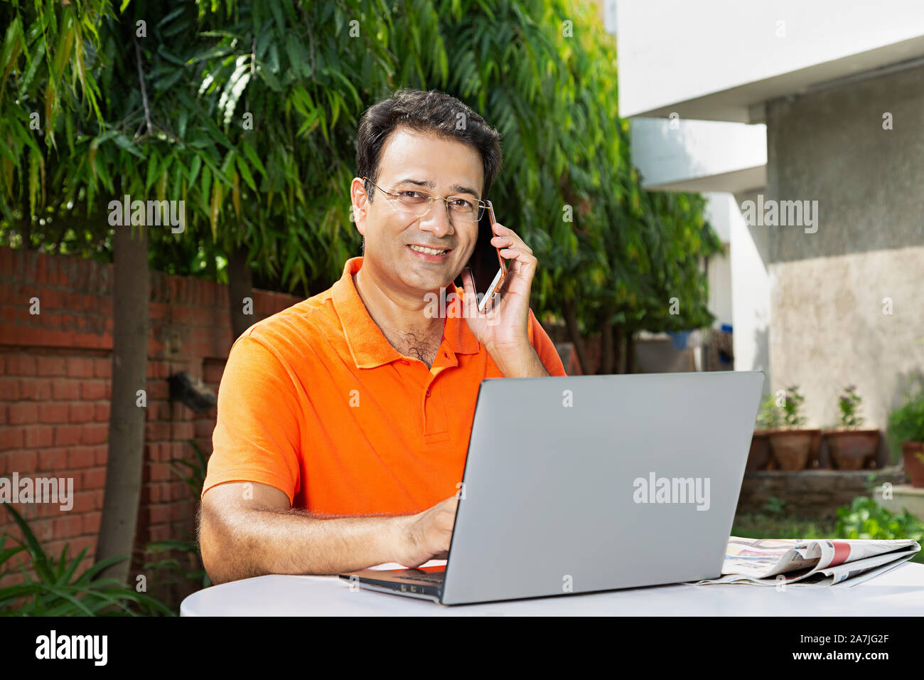 Portrait von lächelnden Mitte erwachsenen männlichen Sitzen-auf-Tabelle Gespräch am Handy mit Arbeiten am Laptop - Hof - Ihr Haus Stockfoto