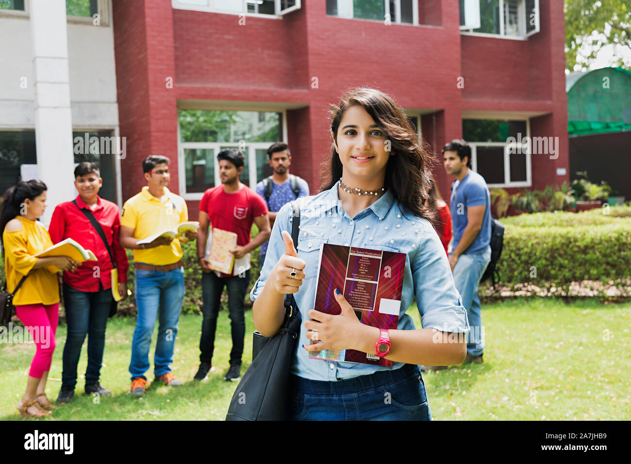 Junge weibliche Studenten Tragetasche angezeigt Thumbs-up mit Buch und Klassenkameraden Im-ohr-Hintergrund außerhalb auf dem Campus Stockfoto