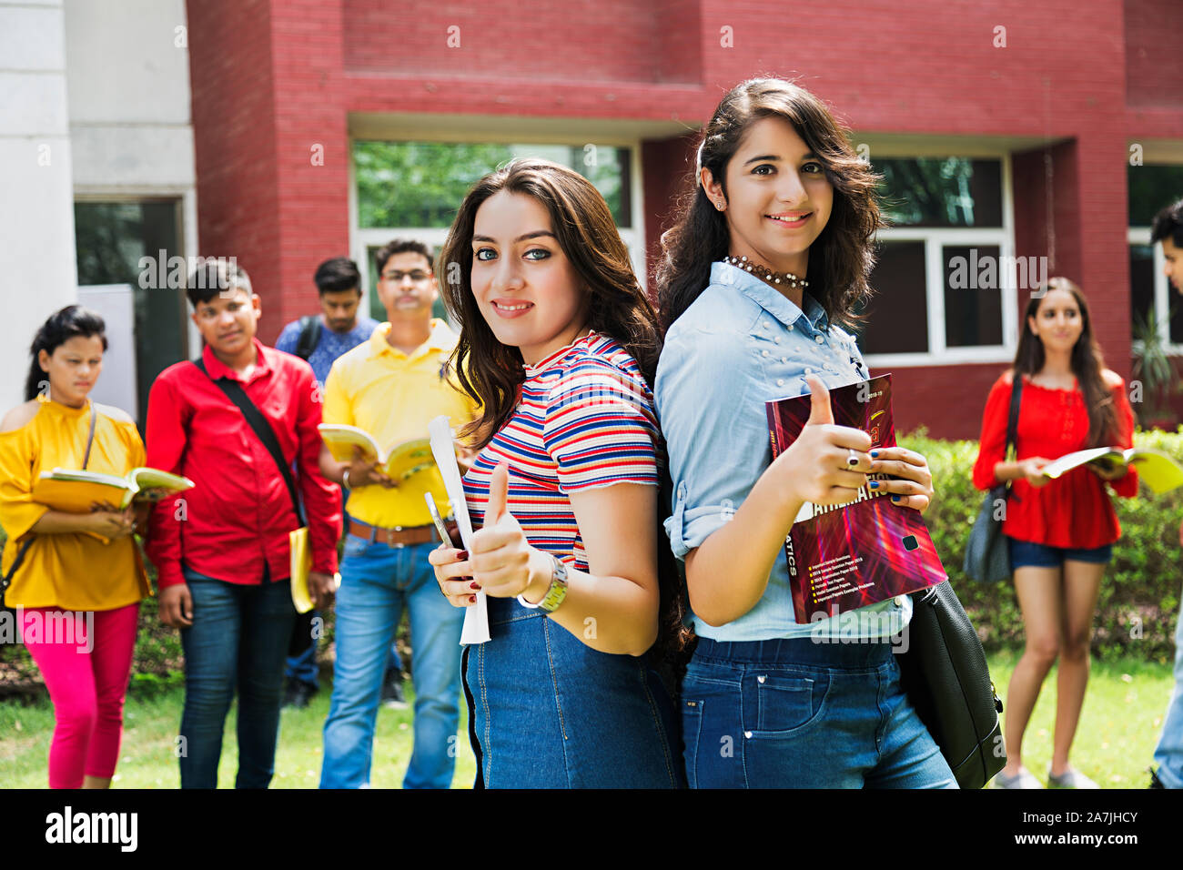 Zwei Teenager Girls College Student Freunde Rücken-an-Rücken und zeigen Thumbs-up mit Classmate-in-Hintergrund - Draußen Campus Stockfoto