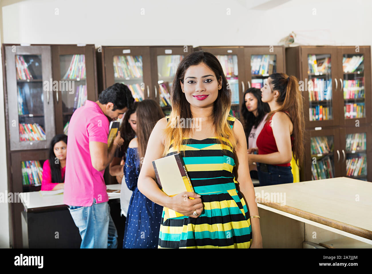 Junge Dame Student Holding Buch Studieren mit Klassenkameraden In-Background At-Library Stockfoto