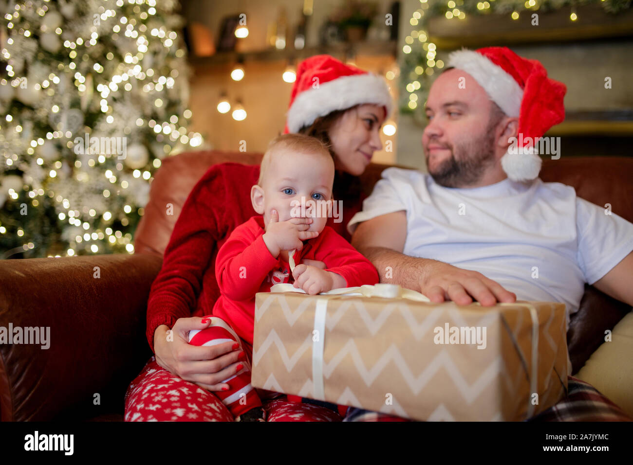 Bild von jungen Eltern in Santa's Caps mit Sohn im Stuhl im Zimmer mit Weihnachtsschmuck Stockfoto