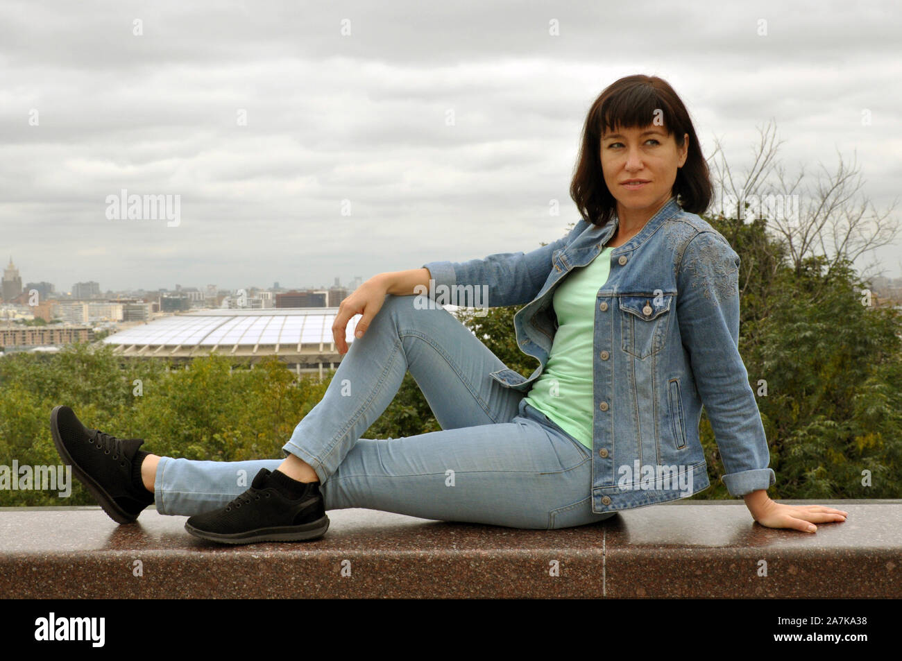 Schöne junge Frau Traveler in Jeans Kleidung sitzt auf der Brüstung und genießt den Blick auf Moskau. Vrorbievy Gory, Blick auf die Stadt Moskau b Stockfoto