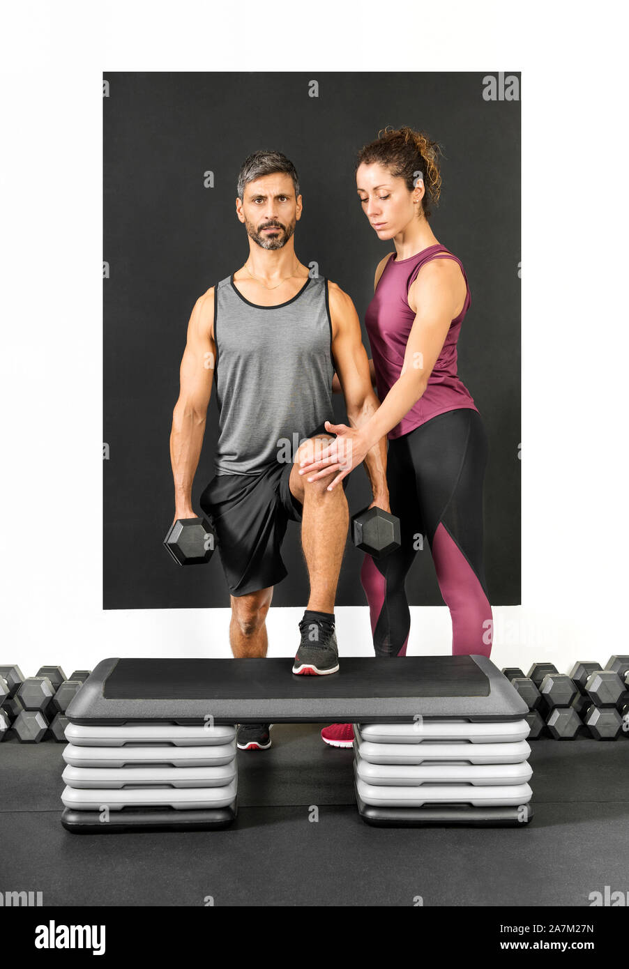 Weibliche personal trainer Unterstützung ein Mann in einer Turnhalle Schritt ups halten zwei kurzhantel Gewichte in einer Gesundheit und Fitness Concept zu tun Stockfoto