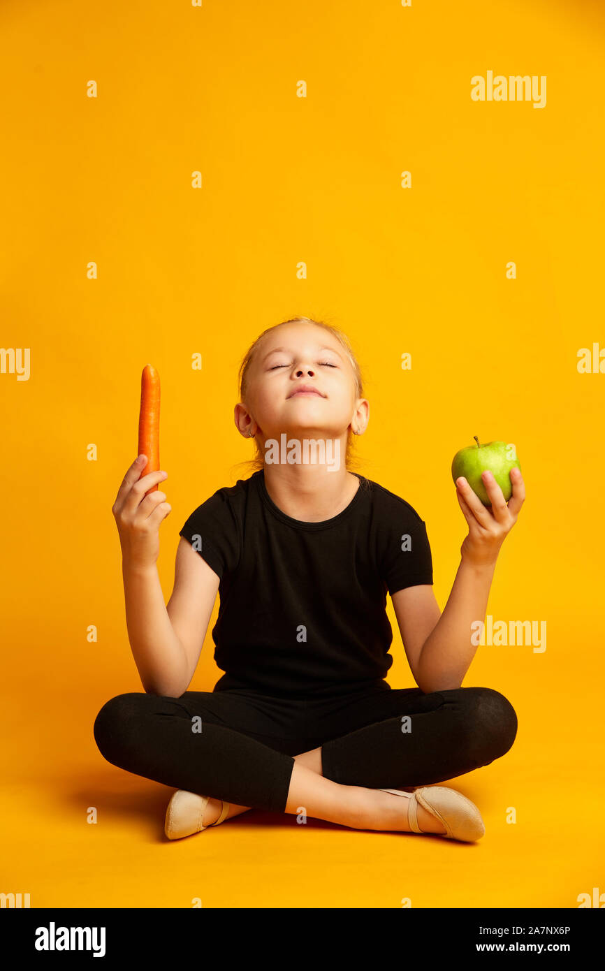 Athletische sieben Jahre alten Mädchen, dass Grüner Apfel und Karotte in der Hand isoliert auf Gelb Stockfoto