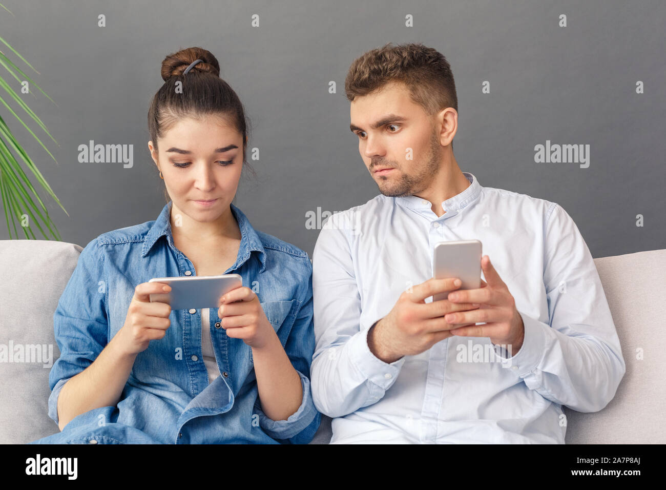 Relationnship Konzept. Junges Paar sitzt auf einem Sofa Studio auf grauen Mann mit Smartphone bei Frau Video Suchen isoliert konzentriert Stockfoto