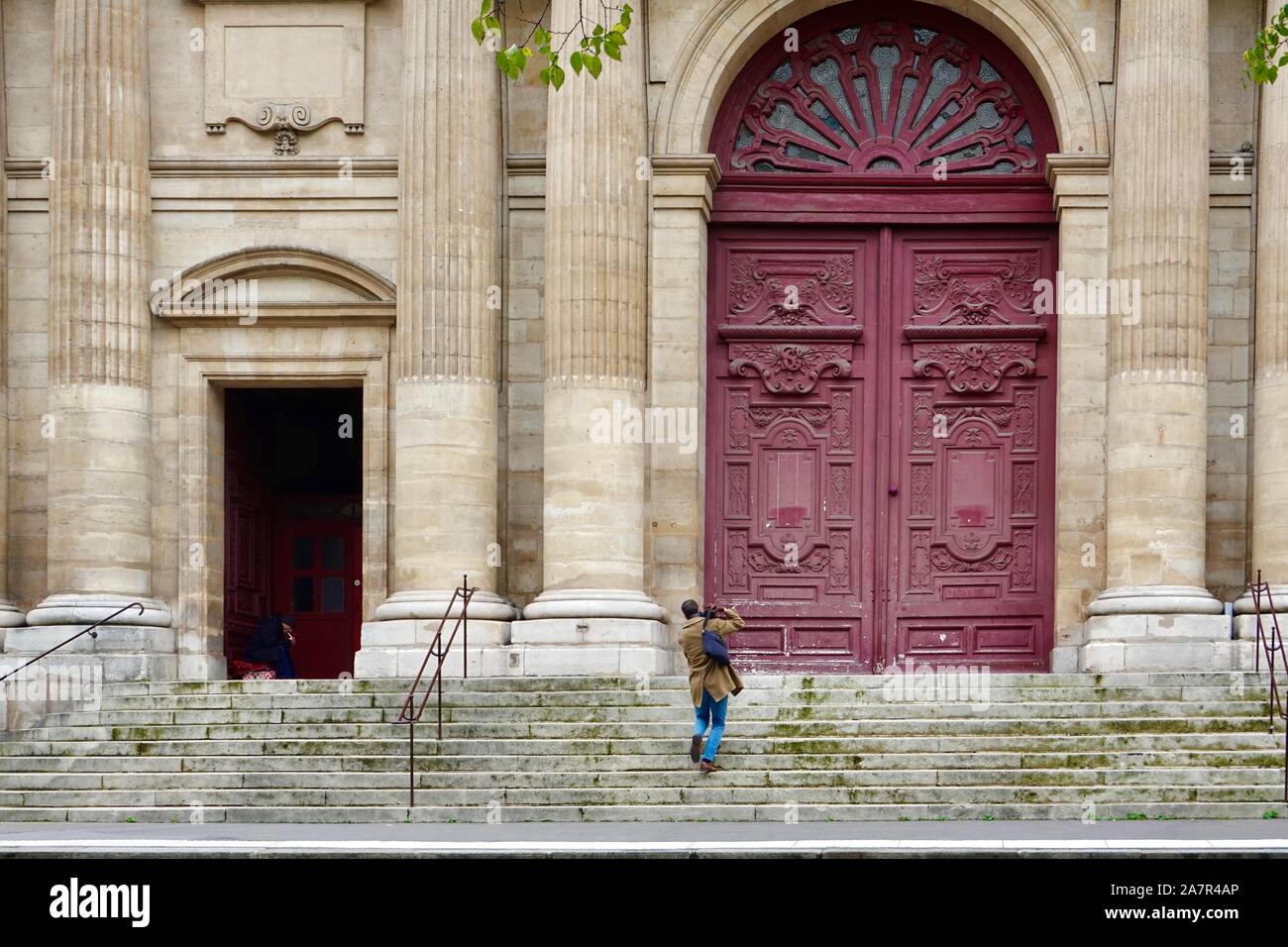 Mann sitzt im Schatten, eine Zigarette, wie ein gut gekleideter Mann in Richtung der Tür der Kirche von Saint-Jean-Baptiste, Paris, FR. Stockfoto