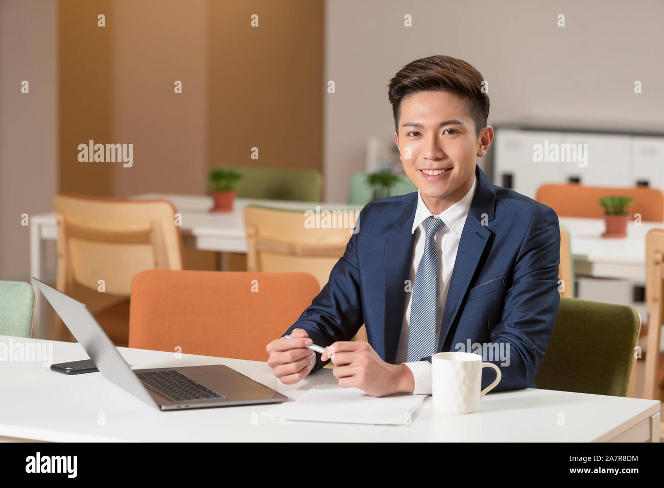 Taille bis Porträt einer jungen lächelnd Geschäftsmann in Anzug sitzen an einem Schreibtisch mit einem Laptop im Büro und lächelnd in die Kamera Stockfoto