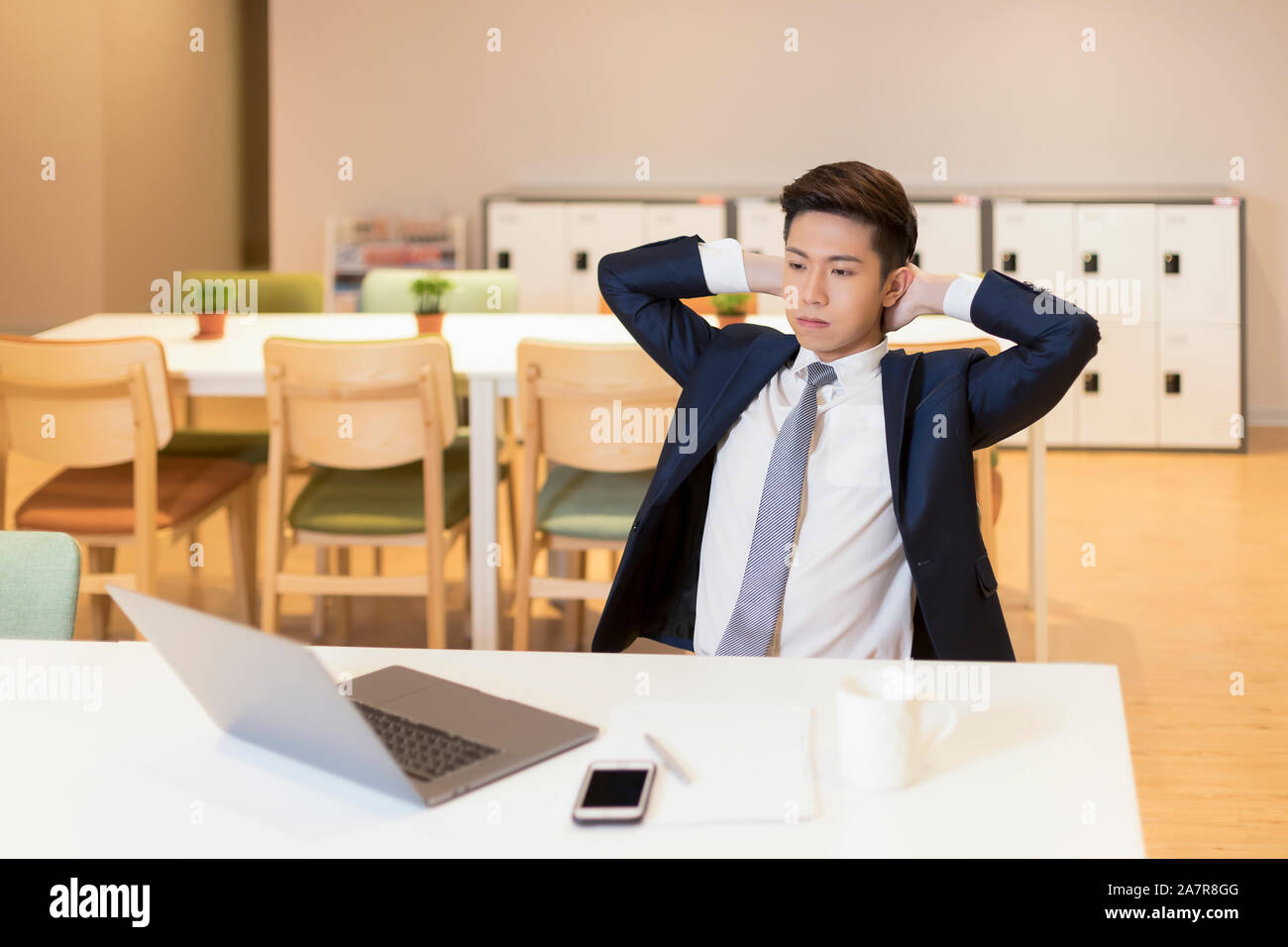 Taille bis Schuß eines junge Unternehmer mit schwarzem Haar und passt auf einen Schreibtisch in einem Büro sitzen mit ernstem Gesichtsausdruck und die Hände hinter den Kopf Stockfoto