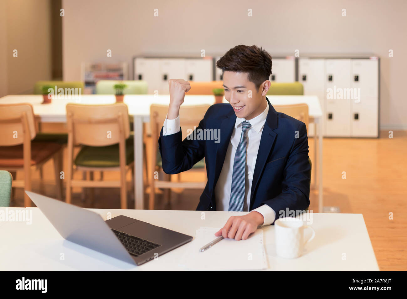 Taille bis Schuß eines junge Unternehmer mit schwarzem Haar und einen Anzug mit einem Erfolg Geste während vor einem Laptop an einem Schreibtisch in einem Büro arbeiten Stockfoto