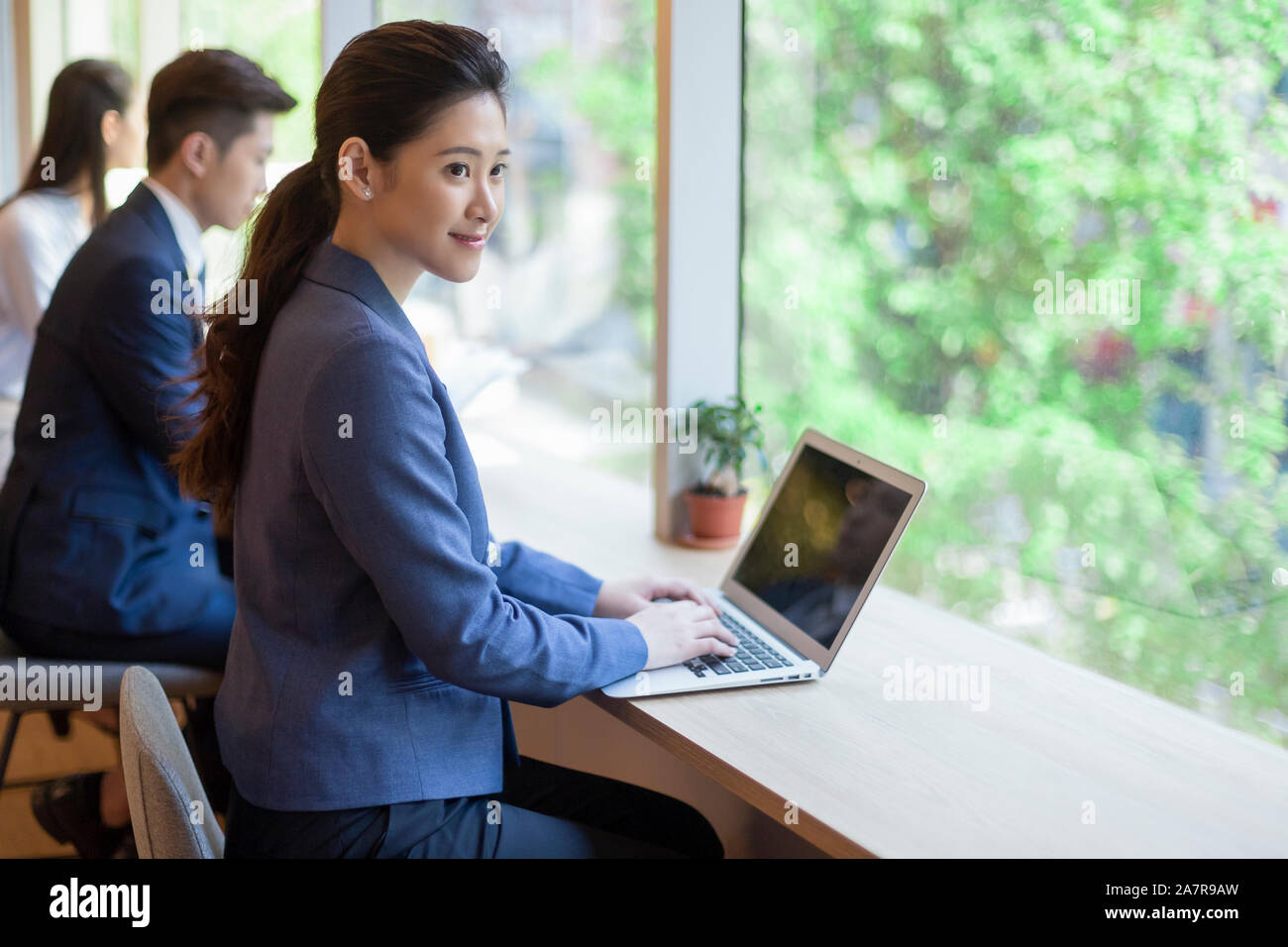 Seitenansicht Schuß eines lächelnden jungen Geschäftsfrau mit einem schwarzen Pferdeschwanz mit einem Laptop an einem Schreibtisch in einem Büro Stockfoto