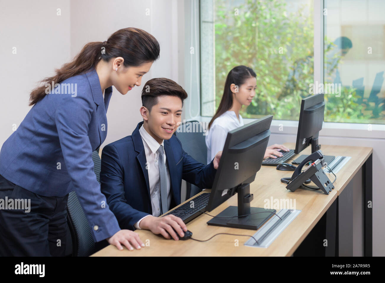 Zwei lächelnde junge männliche und weibliche Unternehmer auf der Suche nach einem Monitor von einem von Ihnen beim zusammen an einem Schreibtisch in einem Büro arbeiten Stockfoto