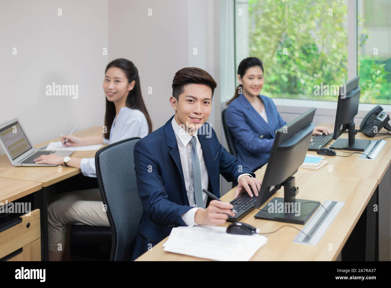 Porträt einer Gruppe von drei lächelnden jungen männlichen und weiblichen Unternehmer sitzen am Schreibtisch in einem Büro und Kamera Stockfoto