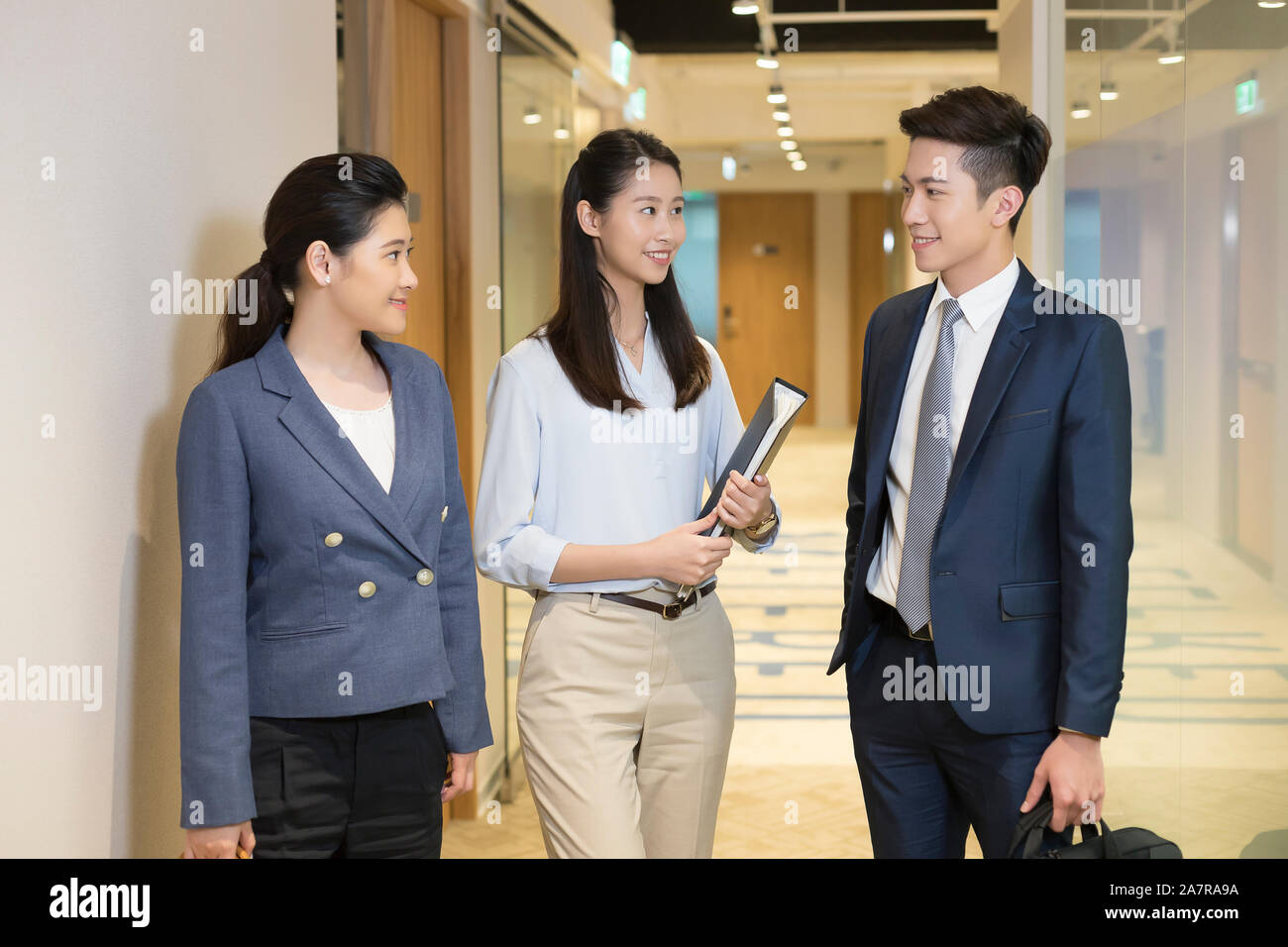 Drei männliche und weibliche junge Geschäftsleute sprechen zusammen und stehen in einem Büro Flur und einer von ihnen ist mit einem Binder Stockfoto