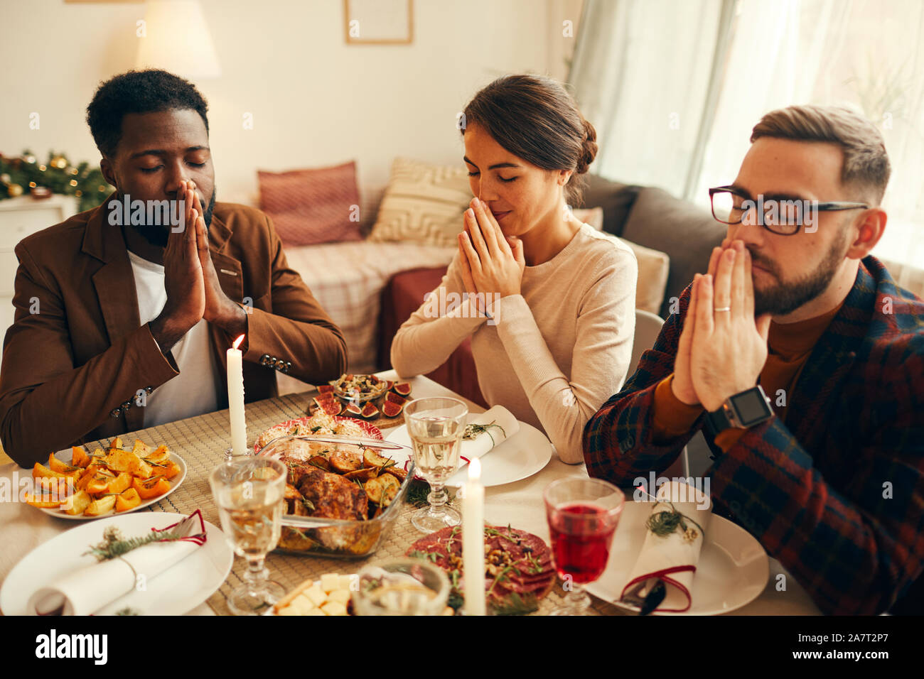 Multi-ethnische Gruppe der elegante erwachsener Menschen, die sagen, dass Gnade Sitzen am Tisch während Weihnachten und Veranstaltungsräume Stockfoto