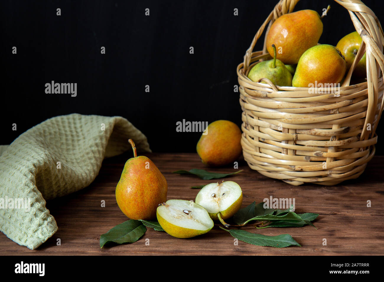 Einen Weidenkorb mit reife Birnen steht auf einem Holztisch. In der Nähe befinden sich drei Birnen. Eine Birne halbiert. Im Hintergrund ist eine grüne Serviette. Stockfoto