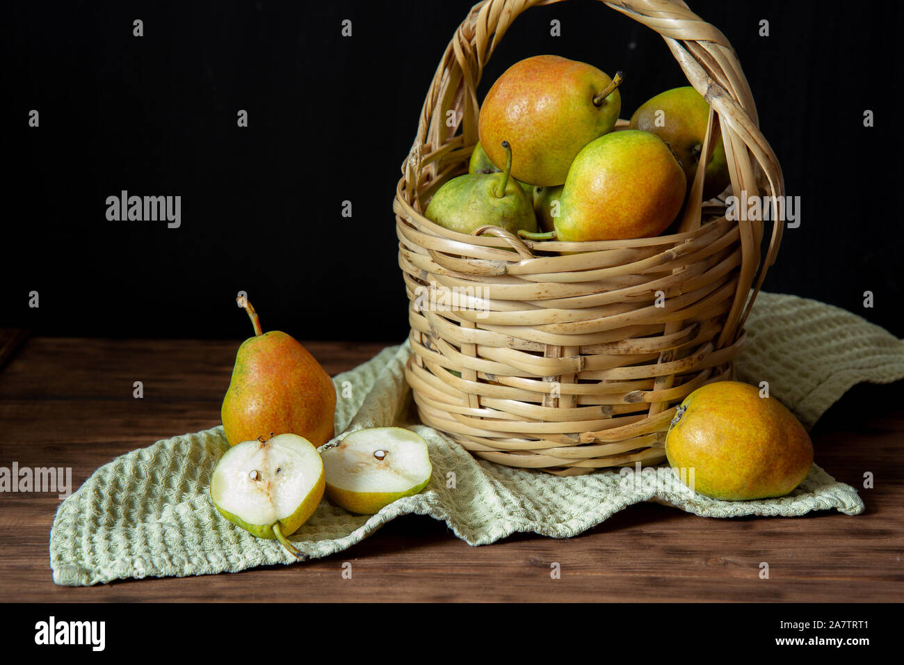 Einen Weidenkorb mit reife Birnen steht auf einem Holztisch. In der Nähe befinden sich drei Birnen. Eine Birne halbiert. Unter dem Korb ist ein grünes Handtuch. Stockfoto