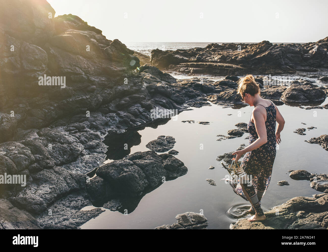 Blonde Frau im Sommerkleid an felsigen Ufer mit Blick auf das Meer in der Sonne Stockfoto