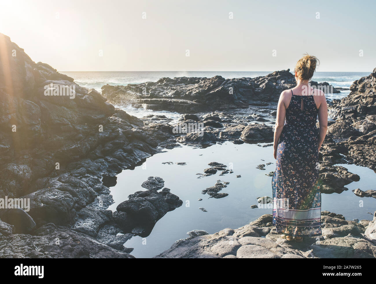 Blonde Frau im Sommerkleid an felsigen Ufer mit Blick auf das Meer in der Sonne Stockfoto