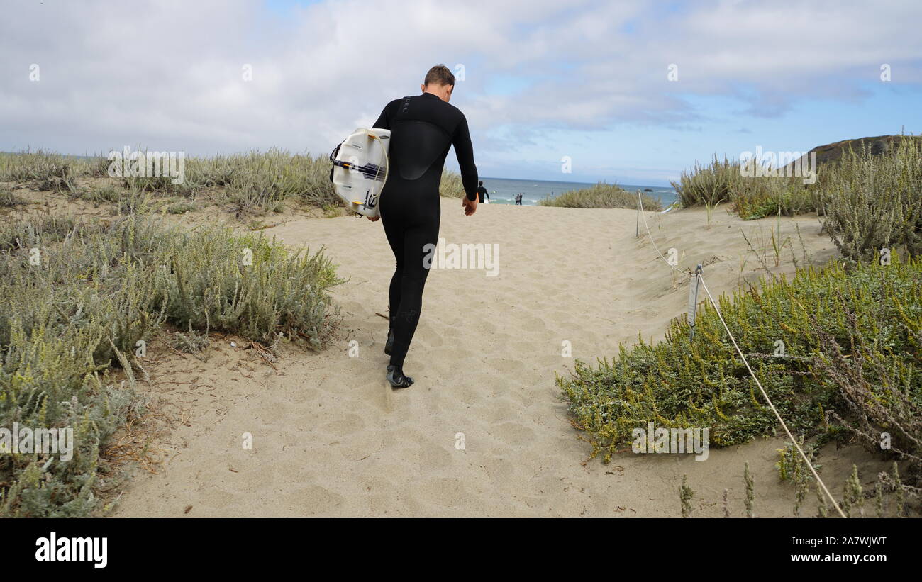 Einen einsamen Mann trägt einen schwarzen Anzug und mit dem Surfbrett ist, ein sandiger Pfad auf der Düne auf den Pazifischen Ozean und bewölkt blauer Himmel Stockfoto