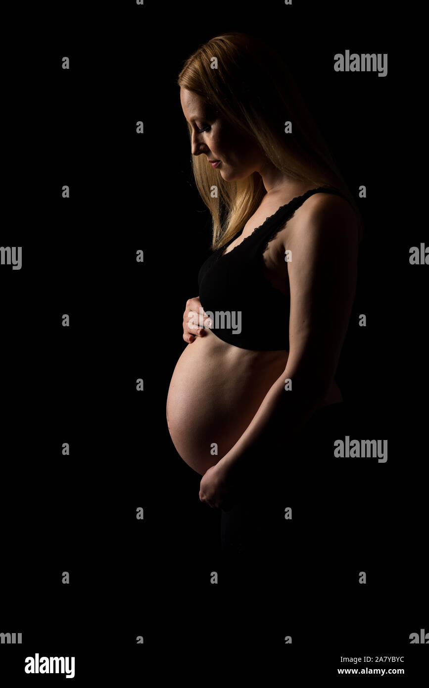 Schwangere Frau in schwarzer Unterwäsche Bauch Holding auf schwarzem Hintergrund. Portrait von aufgeregt, fröhlich, blond, junge Frau in der Schwangerschaft studio schießen. Konzept Stockfoto