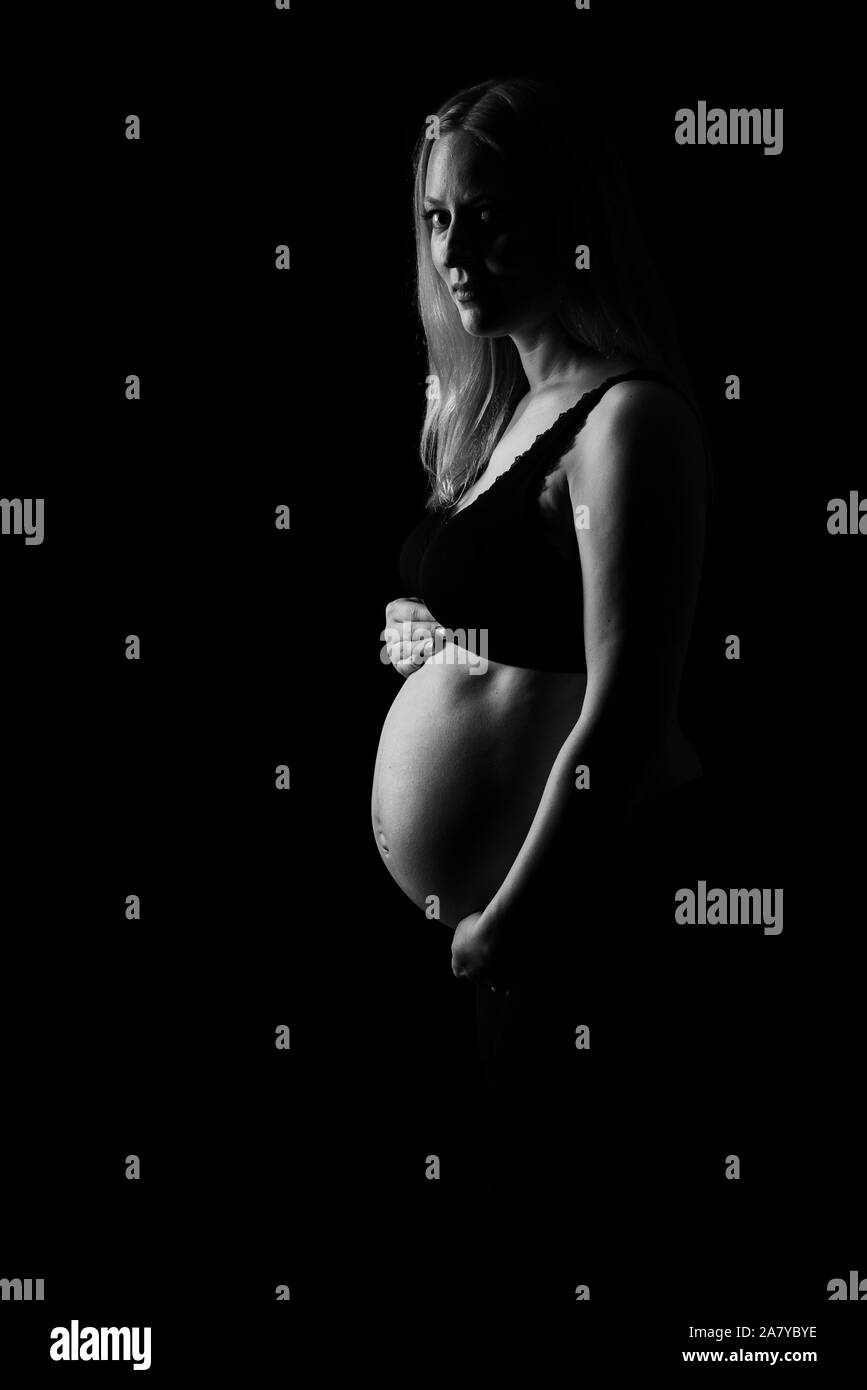 Schwangere Frau in schwarzer Unterwäsche Bauch Holding auf schwarzem Hintergrund. Portrait von aufgeregt, fröhlich, blond, junge Frau in der Schwangerschaft studio schießen. Konzept Stockfoto