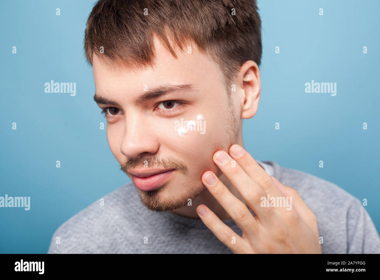 Porträt der jungen brünetten Mann mit dem kleinen Bart und Schnurrbart Anwendung Gesichtscreme unter Augen, Behandlung oder Feuchtigkeitscreme Kosmetik, Hautpflege Konzept. Ich Stockfoto