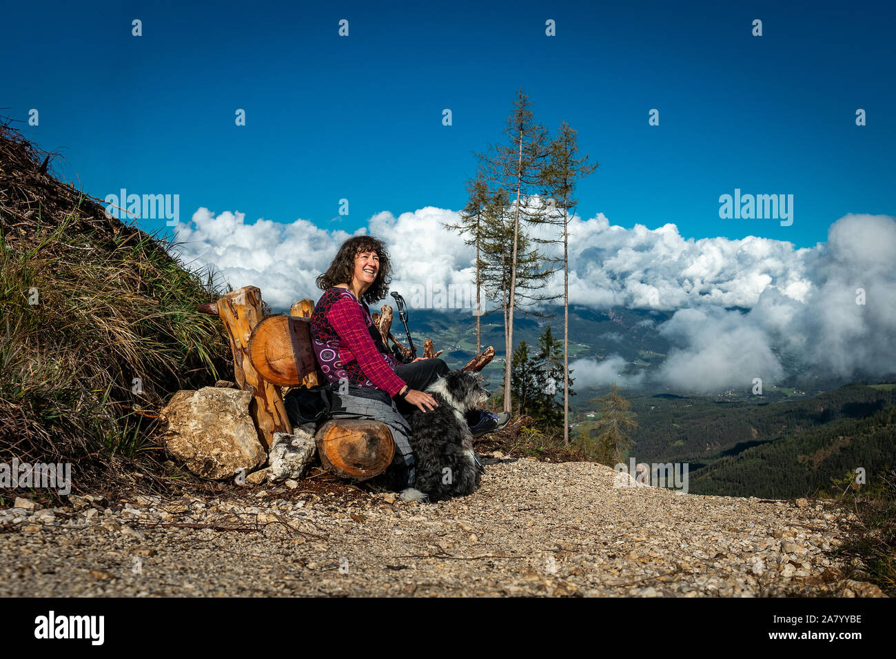 Frau und Hund sitzen auf einer Bank ausruhen beim Wandern zu den Tschaffonhuette auf einem hohen Berg in der italienischen Schlern Dolomitengebiet mit weißen Wolken Stockfoto