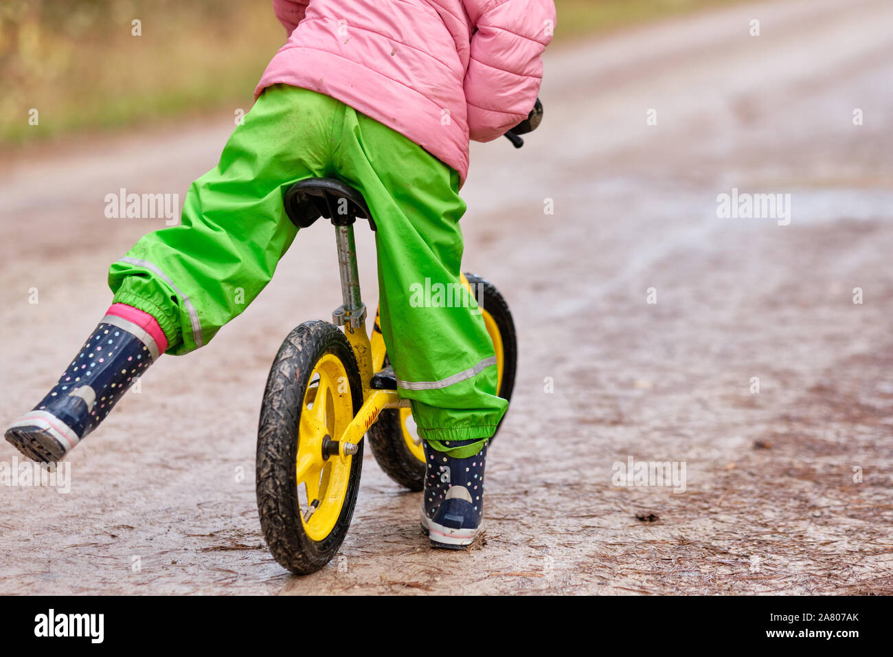 Ansicht der Rückseite des unteren Bereich eines Kindes Mädchen erhalten auf ihre Balance Fahrrad in warme und wasserdichte Kleidung auf einem feuchten und verschmutzten Schotterstraße im Herbst. Gesehen Stockfoto