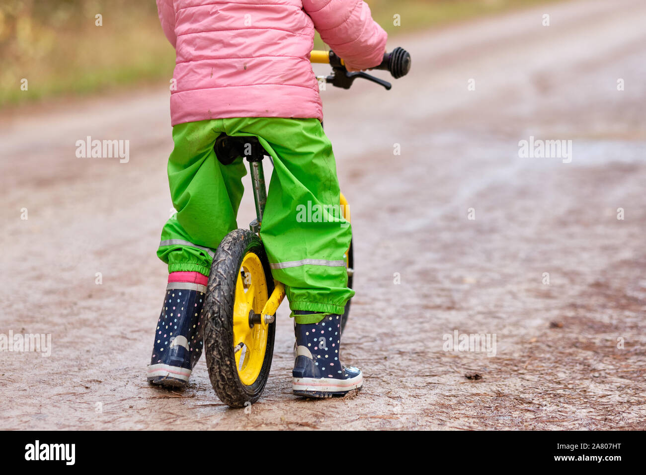 Ansicht der Rückseite des unteren Bereich eines Kindes Mädchen sitzen auf ihr Gleichgewicht Fahrrad in warme und wasserdichte Kleidung auf einem feuchten und verschmutzten Schotterstraße im Herbst. Gesehen Stockfoto