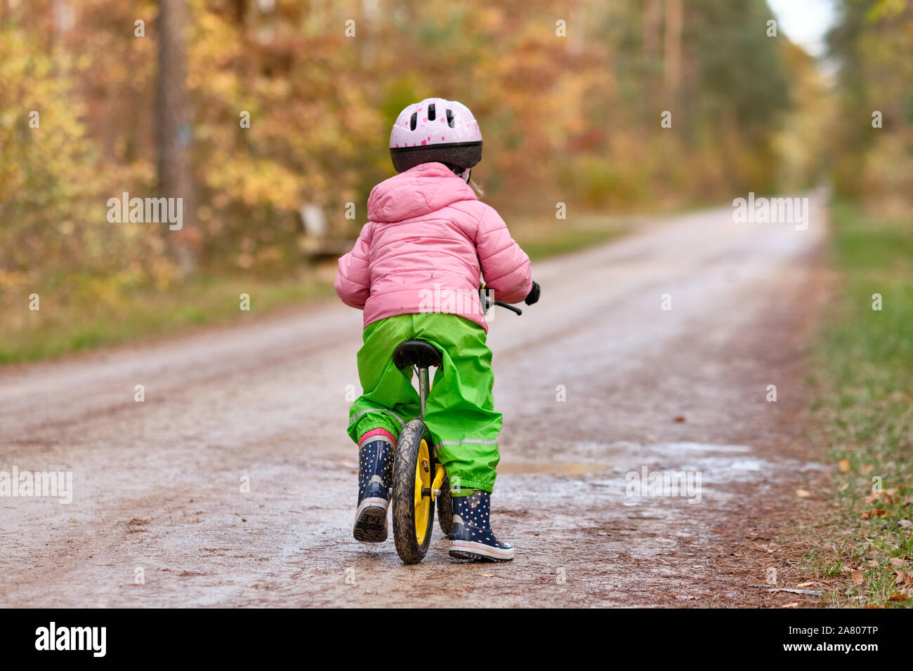 Rückansicht eines Kindes Mädchen reiten auf ihre Balance Fahrrad in warme und wasserdichte Kleidung auf einem feuchten und verschmutzten Schotterweg in den Wald. In F gesehen Stockfoto