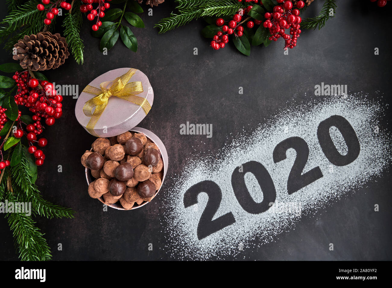 Wir begrüßen das Jahr 2020 mit einer Schachtel Pralinen und Schokolade Trüffel, einige Ananas und einige holly Filialen auf einem dunklen Hintergrund. Stockfoto