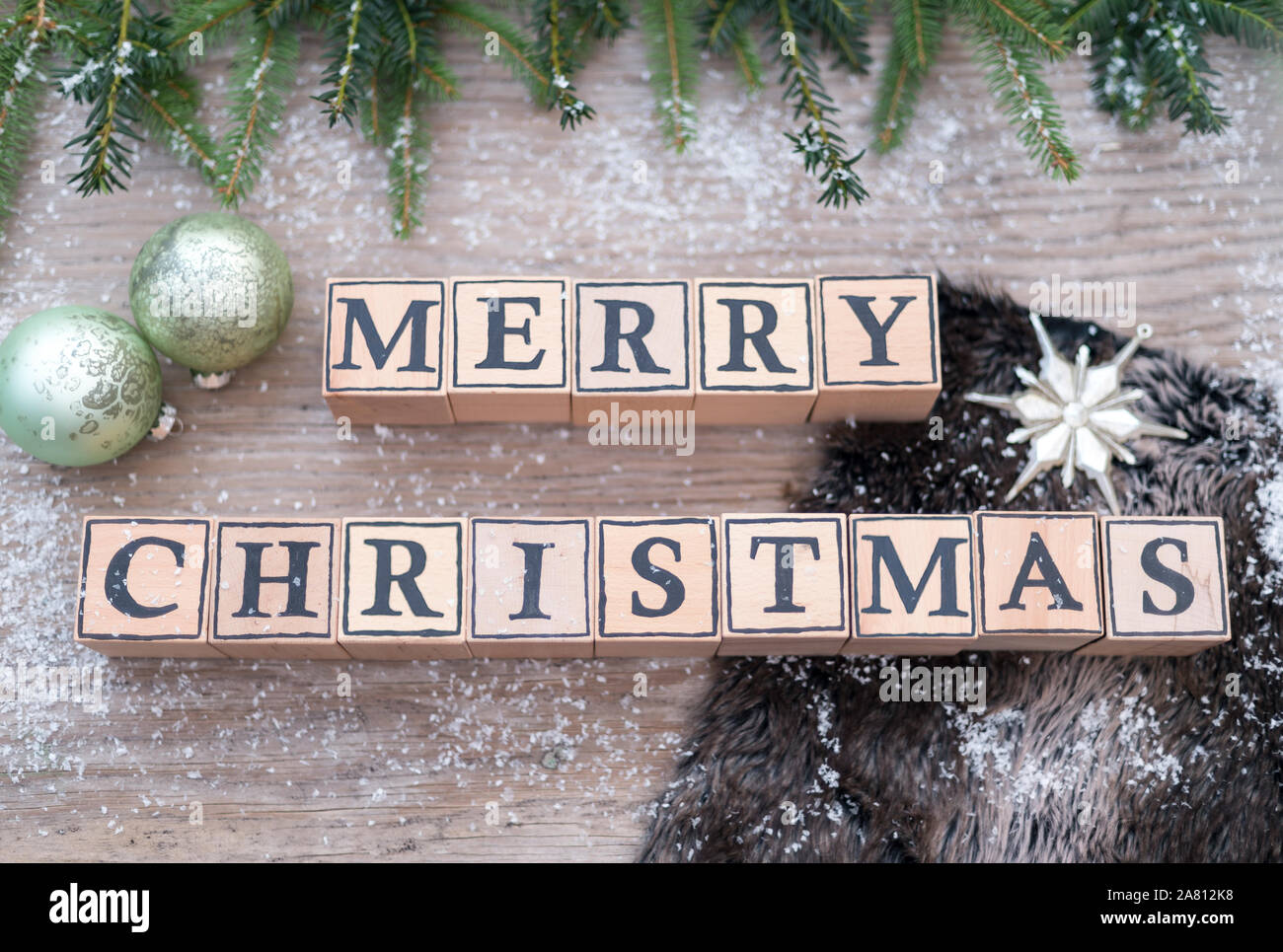 Holzwürfel mit Buchstaben Form der englische Text frohe Weihnachten. Sie liegen auf einem Holz Hintergrund. Tannenzweigen und Weihnachten Kugeln sind. Stockfoto