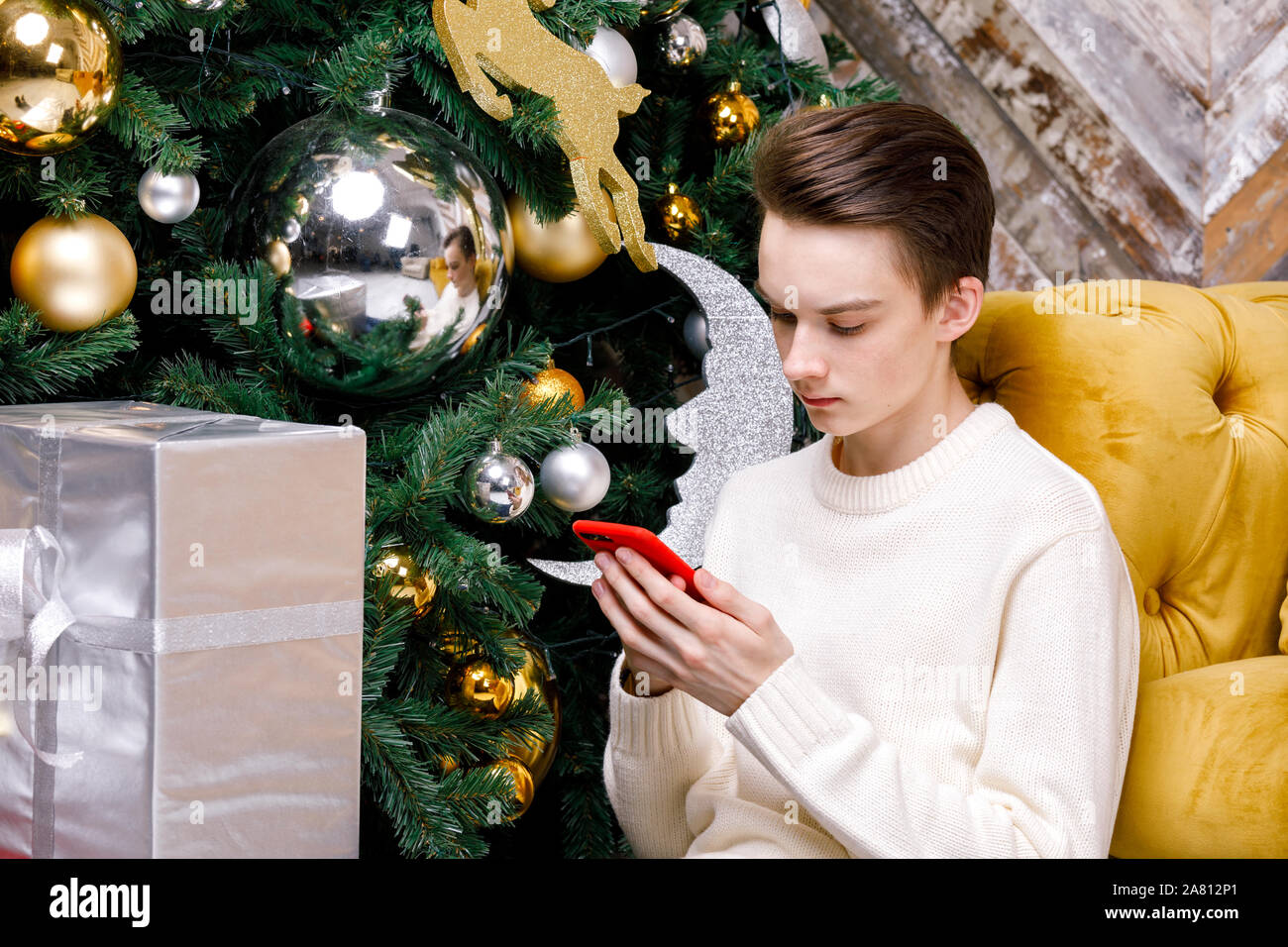 Teenager Junge allein sitzen auf einem Fußboden durch Weihnachtsbaum mit Geschenken konzentrierte sich nur auf sein Smartphone Messaging oder Kontrolle soziale Medien - Smombie Urlaub Stockfoto