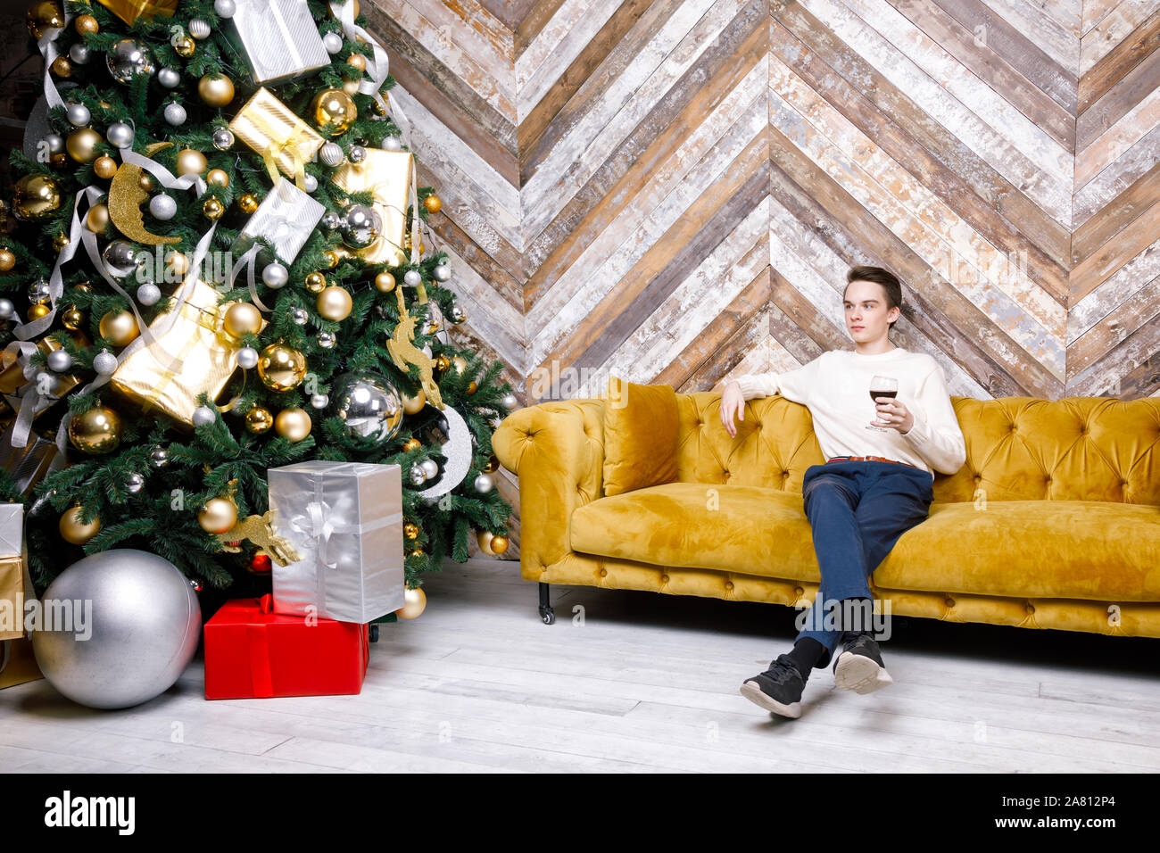 Teenager Junge allein Sitzen auf einem Sofa neben dem Weihnachtsbaum mit Geschenken holding Glas Rotwein feiern Weihnachten Urlaub - teenage Alkohol Konsum Stockfoto