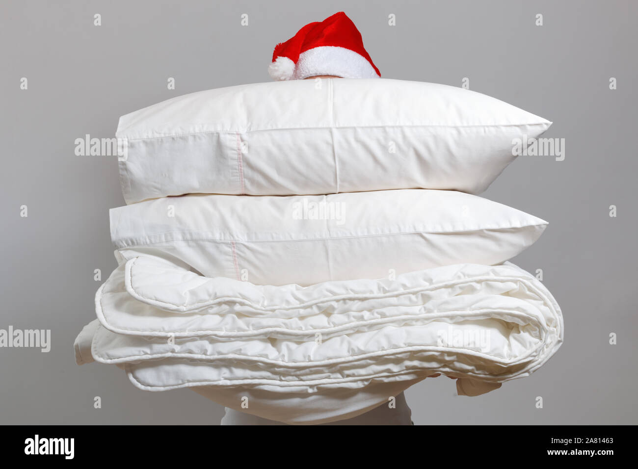 Frau in santa hut mit einem warmen Federbett und Federkissen gegen eine graue Wand. Stapel von Betten zum Schlafen. Haushalt zu Weihnachten. Stockfoto