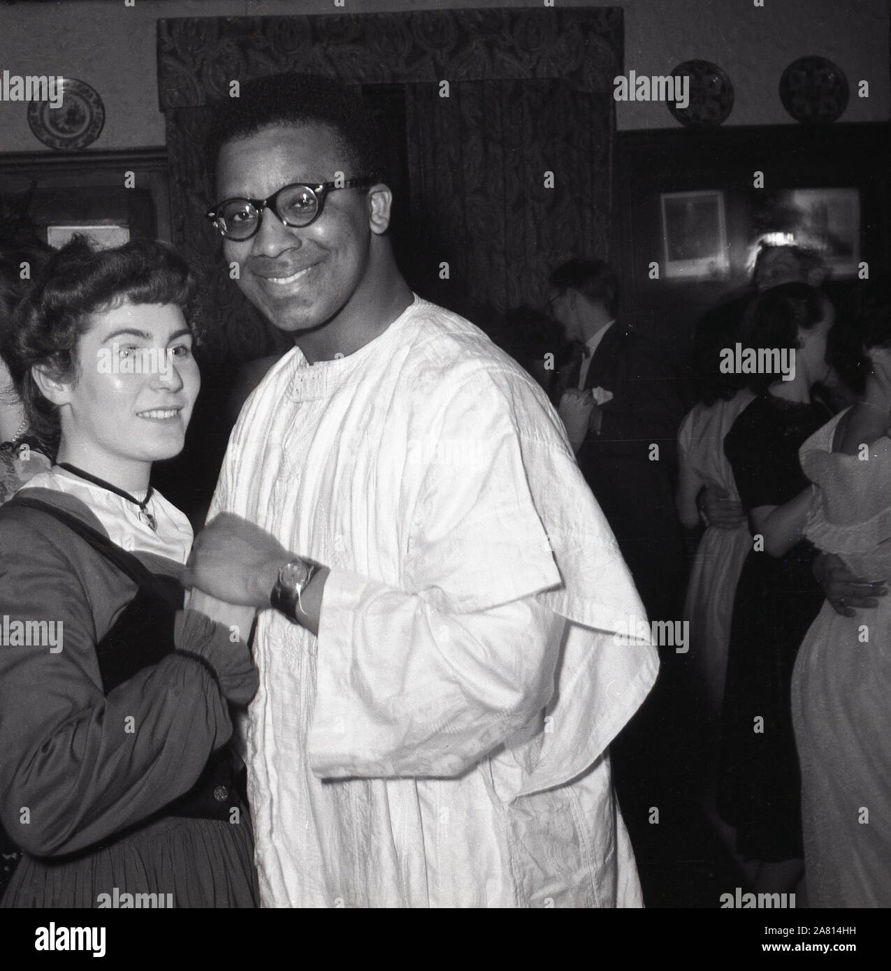 1950er Jahre, historisches, Nachkriegs-Großbritannien und eine junge englische Dame, die mit einem Überseemänner aus Westafrika tanzt, der ein traditionelles, lang fließendes Gewand trägt, eine Agbada, die normalerweise von Männern in Nigeria und anderen westafrikanischen Ländern, London, England, Großbritannien getragen wird. In dieser Ära kamen viele Völker aus den überseeischen Commonwealth-Ländern nach Großbritannien, um zu arbeiten und zu studieren und Funktionen und Tänze zu spielen und wurden gehalten, um sie in die britische Lebensweise einzuführen. Stockfoto