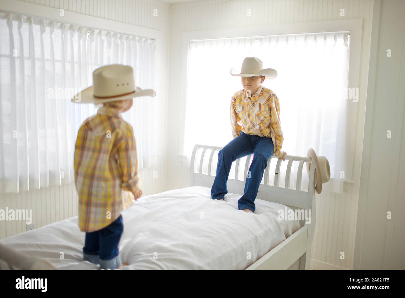 Die beiden jungen Bruder tragen Cowboyhüte, stehend auf einem Bett aus Holz in einem sonnigen weißen Raum. Stockfoto