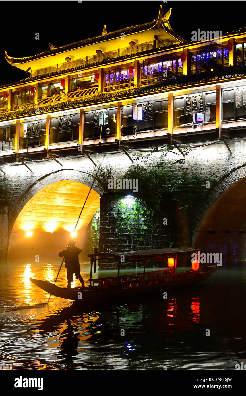 Die nächtliche Silhouette eines Mannes mit einem Sampan Boot schwimmt unter den Bögen der hell erleuchteten Phoenix Hong Brücke in Fenghuang antike Stadt in Stockfoto