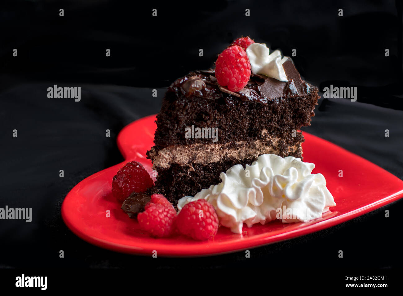 Ein schönes Stück Schokoladenkuchen, beruht auf einer roten Herzen Platte und ist mit Schlagsahne garniert, Himbeeren und rasiert Schokolade Stockfoto