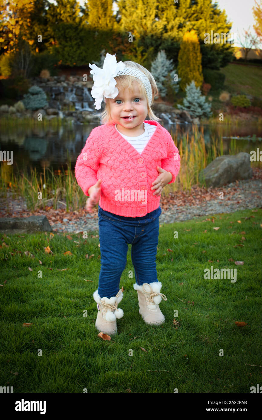 Sehr niedliches Kleinkind blonde Mädchen mit großen blauen Augen lächeln Mitten im schönen Herbst Park tragen Rosa Jacke und blaue Jeans und Stockfoto