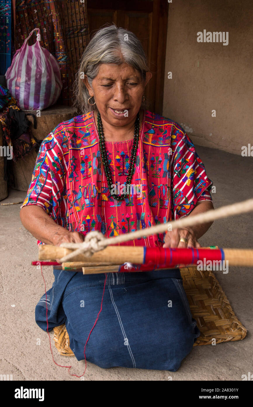 Eine ältere, grauhaarige Frau Maya webt Stoff auf einem Backstrap Loom kniend auf dem Boden ihres Hauses in Santa Catarina Palopo, Guatemala. Stockfoto