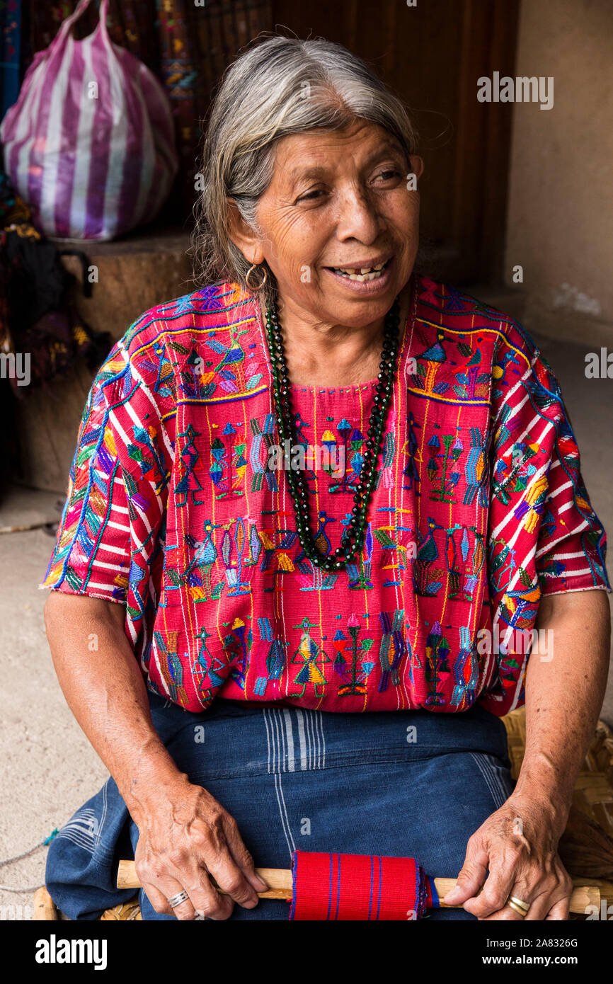 Eine ältere, grauhaarige Frau Maya webt Stoff auf einem Backstrap Loom kniend auf dem Boden ihres Hauses in Santa Catarina Palopo, Guatemala. Stockfoto