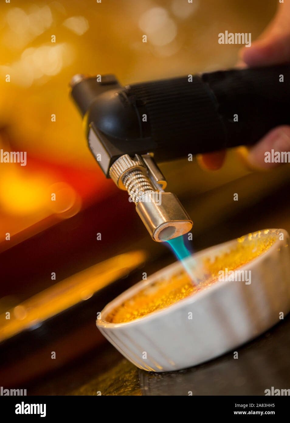 Creme Brûlée Fackel beleuchtete und karamelisierte die Spitze des Desserts mit Bokeh im Hintergrund Stockfoto