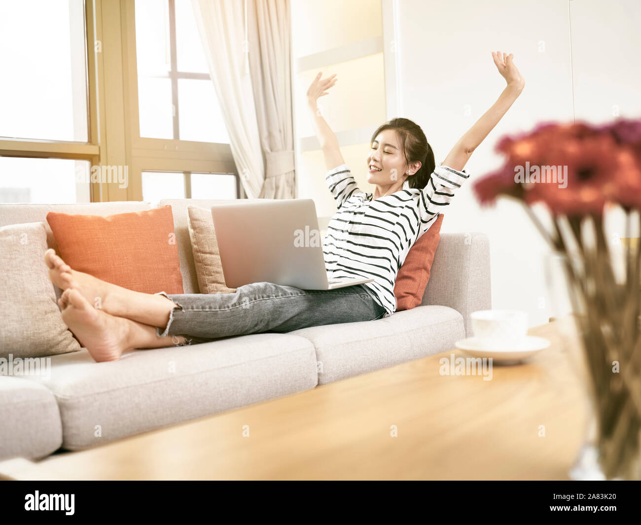 Schöne und glückliche junge asiatische Frau von zu Hause aus arbeiten Sittng auf mit laptop computer Couch, erhobenen Armen Erfolg feiern. Stockfoto