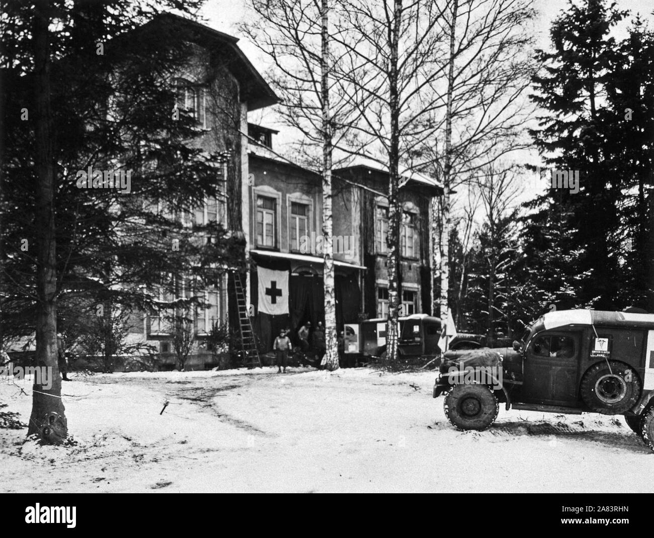 Vorne Außenansicht eines Landes Villa, die in ein medizinisches clearing Station umgewandelt wurde. Schnee bedeckt die vordere Hof. Zwei US-Armee-Lkw sitzen vor. Die hintere Tür des Fahrzeugs am nächsten an der Villa wird geöffnet, und Militärs Schweben zwischen dem Fahrzeug und dem Gebäude. Stockfoto